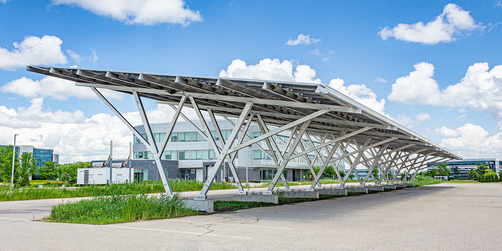 Dramatic photo of an aluminum solar carport at midday. Sky is fair-weather with white clouds. Shot in a parking lot with lush grass verges in the background
